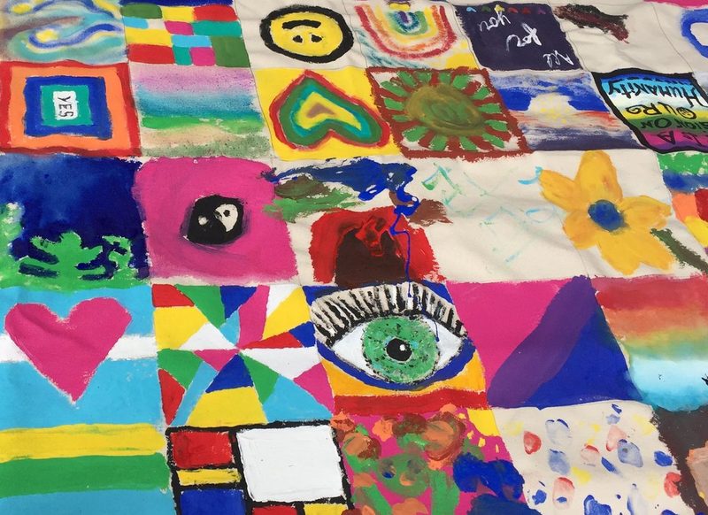 A long canvas of small painted squares, each one made by a different person, bright with rainbows, hearts, an eye and the word yes